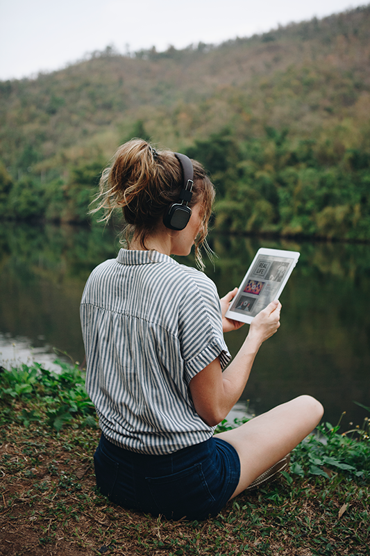 Woman wearing headphones sitting along the river bank looking at a tablet